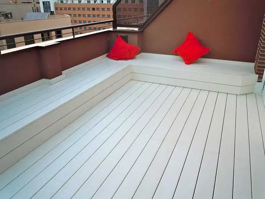 Tarima de composite en terraza Tarima de composite clara en una terraza, con banco integrado y cojines rojos